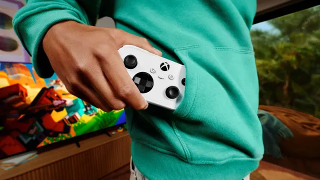 A hand pulling a white Xbox Nano Controller out of a green hoodie pocket, with Minecraft on a TV in the background