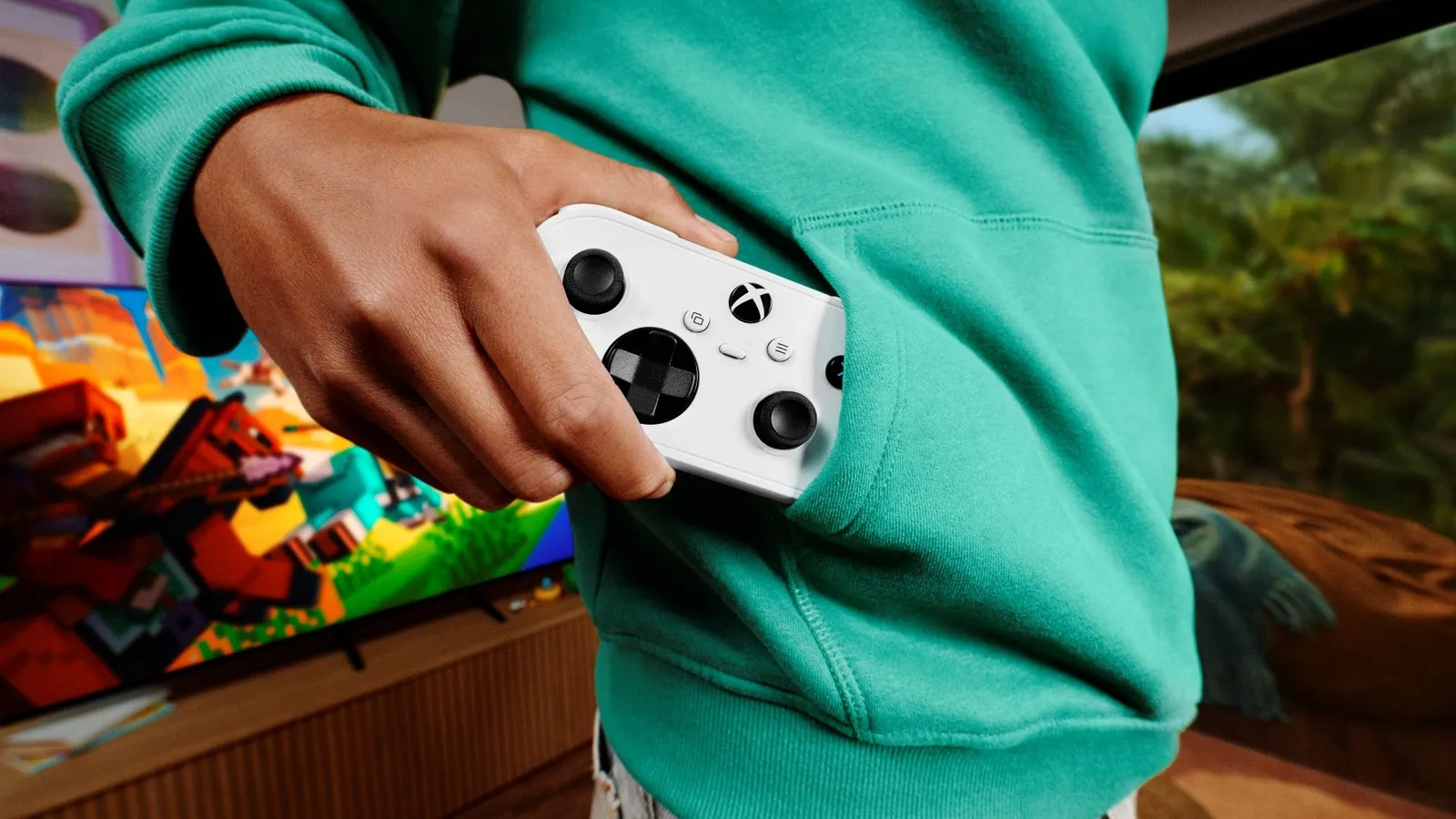 A hand pulling a white Xbox Nano Controller out of a green hoodie pocket, with Minecraft on a TV in the background